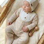 نوزاد با سرهمی بژ و کلاه سفید در سبد حصیری، عکس تنخور با نور طبیعی کنار پنجره