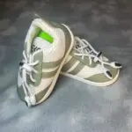کفش کتانی نوزادی زیتونی و کرم با رویه توری، سه نوار کناری و بند کشی قفل‌دار روی پس‌زمینه طوسی